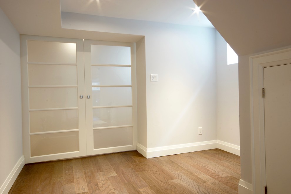 Medium sized contemporary fully buried basement in Toronto with white walls, medium hardwood flooring and beige floors.