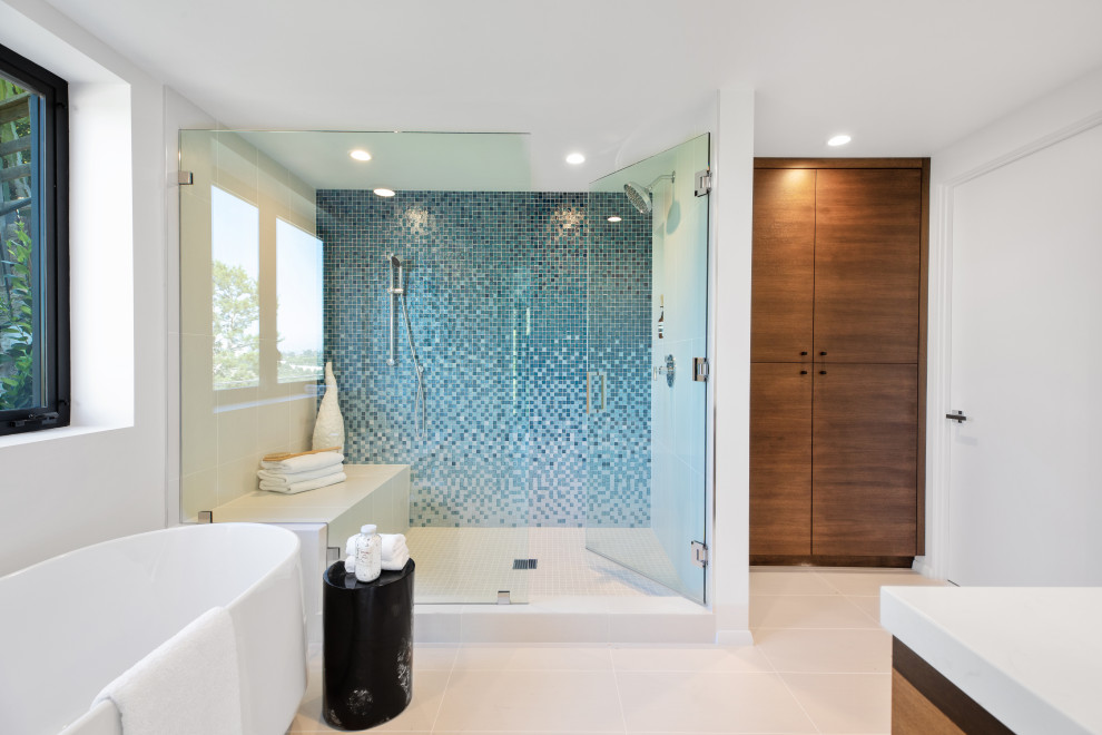 Modernes Badezimmer mit hellbraunen Holzschränken, freistehender Badewanne, Duschnische, blauen Fliesen, Mosaikfliesen, weißer Wandfarbe, grauem Boden, Falttür-Duschabtrennung, weißer Waschtischplatte und Duschbank in Los Angeles