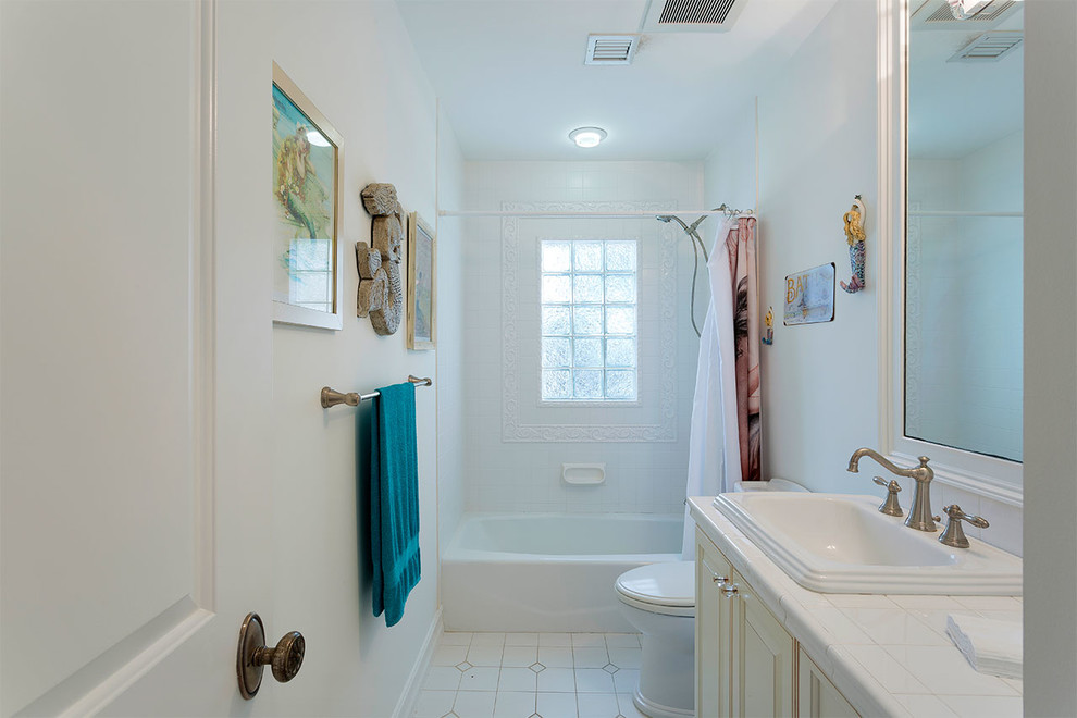 Cette image montre une salle de bain principale marine de taille moyenne avec un placard avec porte à panneau surélevé, des portes de placard beiges, une baignoire en alcôve, un combiné douche/baignoire, WC à poser, un carrelage blanc, des carreaux de porcelaine, un mur blanc, un sol en carrelage de terre cuite, un lavabo posé, un plan de toilette en carrelage, un sol blanc, une cabine de douche avec un rideau et un plan de toilette blanc.