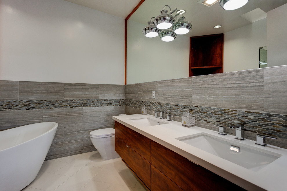 Cette photo montre une grande salle de bain principale moderne en bois brun avec un placard à porte plane, une baignoire indépendante, WC à poser, un carrelage gris, un carrelage en pâte de verre, un mur blanc, un sol en carrelage de céramique, un lavabo intégré et un sol beige.