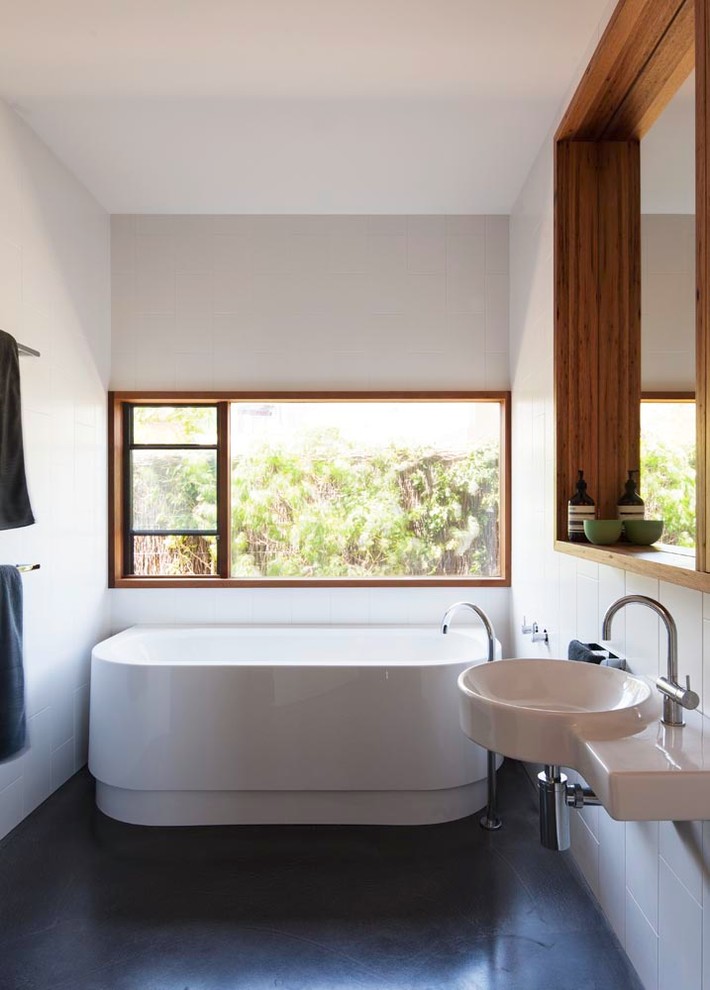 Foto på ett mellanstort funkis badrum, med ett väggmonterat handfat, skåp i ljust trä, ett fristående badkar, en dusch i en alkov, vit kakel, keramikplattor, vita väggar och betonggolv