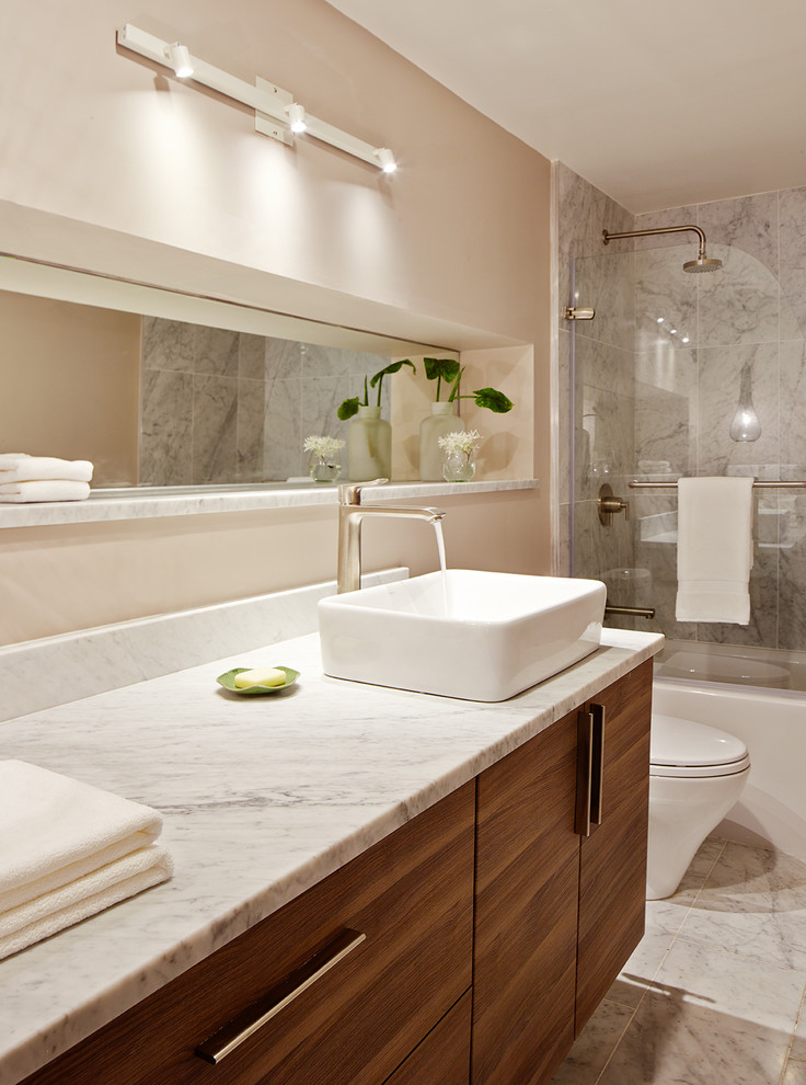 Modern inredning av ett mellanstort en-suite badrum, med ett fristående handfat, släta luckor, skåp i mörkt trä, marmorbänkskiva, ett badkar i en alkov, en dusch/badkar-kombination, en toalettstol med hel cisternkåpa, stenkakel, beige väggar, marmorgolv och vit kakel