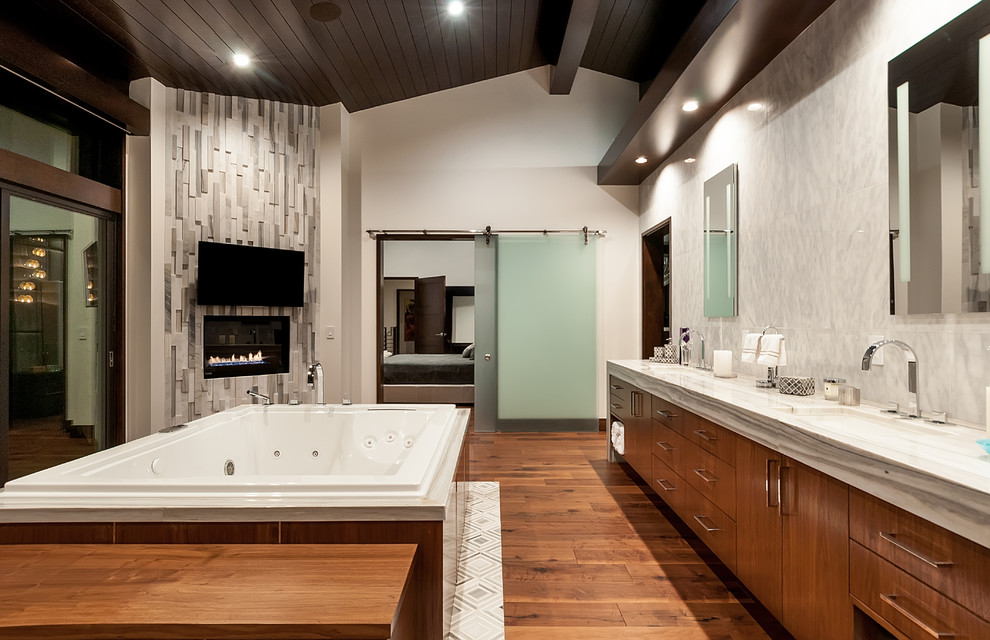 Cette photo montre une salle de bain principale tendance en bois brun avec un placard à porte plane, un bain bouillonnant, un carrelage gris, un mur gris, un sol en bois brun, un lavabo encastré et un sol marron.