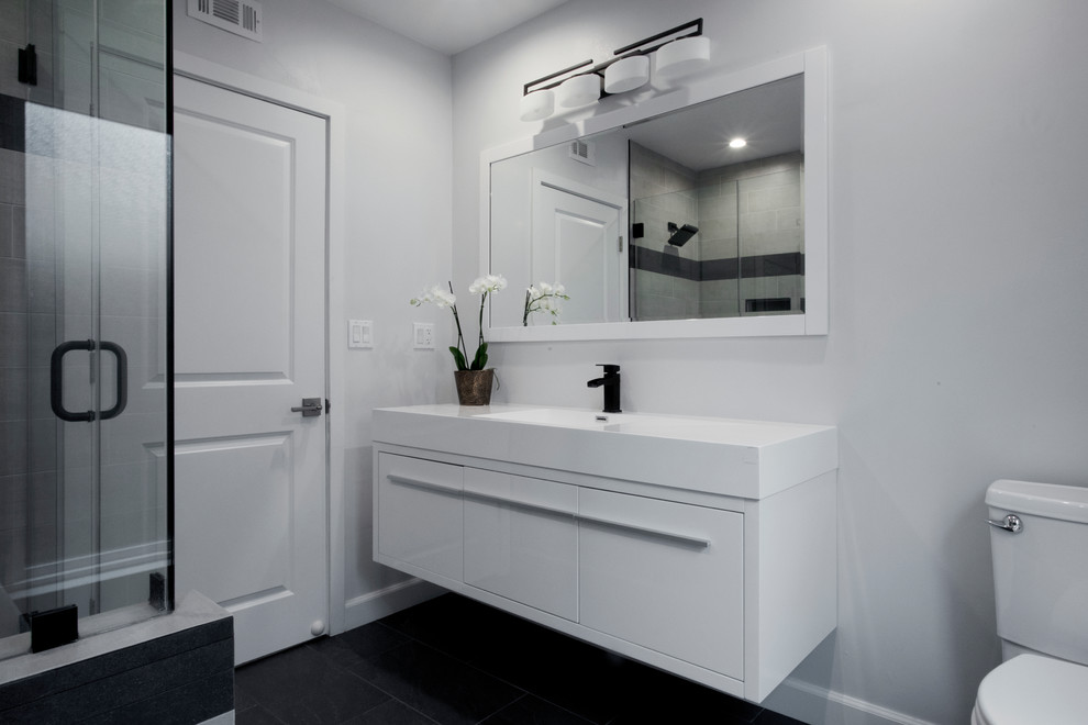 На фото: детская ванная комната среднего размера в стиле модернизм с плоскими фасадами, белыми фасадами, ванной в нише, открытым душем, унитазом-моноблоком, серой плиткой, керамогранитной плиткой, белыми стенами, полом из сланца, монолитной раковиной, столешницей из искусственного камня, черным полом, душем с распашными дверями и белой столешницей с