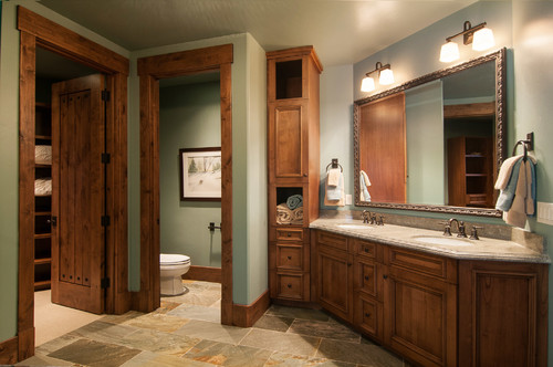 Rustic bathroom cabinets in knotty wood with an L-shaped vanity, stone countertop, tall tower storage, framed mirror, slate floor, and bronze faucets under sconces.