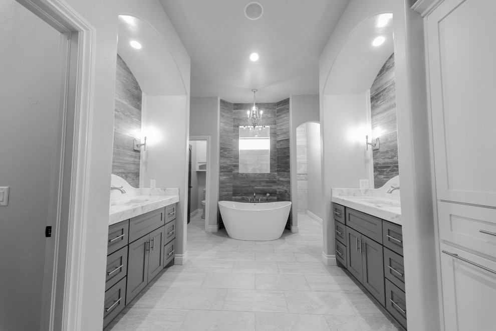 Exemple d'une grande salle de bain principale chic avec un placard à porte shaker, des portes de placard grises, une baignoire indépendante, un combiné douche/baignoire, WC séparés, un carrelage beige, un carrelage de pierre, un mur gris, un sol en carrelage de porcelaine, un lavabo encastré et un plan de toilette en marbre.