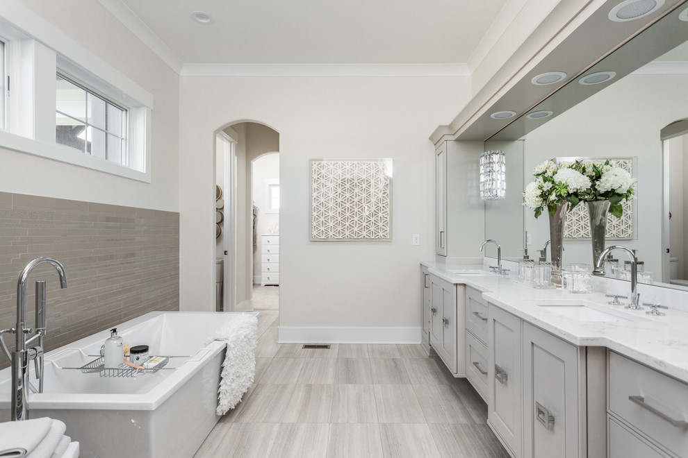 Aménagement d'une grande salle de bain principale contemporaine avec un placard avec porte à panneau encastré, des portes de placard grises, une baignoire indépendante, un carrelage gris, un mur beige, un lavabo encastré, un sol en carrelage de porcelaine, un plan de toilette en marbre, un sol beige et une fenêtre.