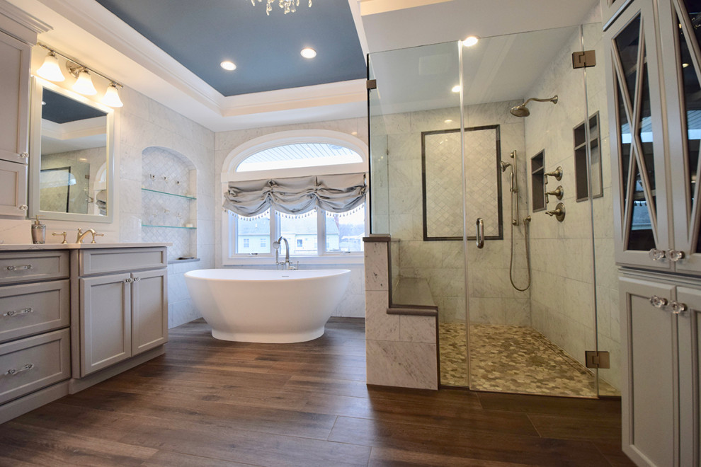 Свежая идея для дизайна: большая главная ванная комната в классическом стиле с фасадами с утопленной филенкой, серыми фасадами, отдельно стоящей ванной, душем без бортиков, унитазом-моноблоком, белой плиткой, серыми стенами, полом из керамогранита, врезной раковиной, столешницей из искусственного кварца, коричневым полом, душем с распашными дверями, белой столешницей, сиденьем для душа, тумбой под две раковины, напольной тумбой, многоуровневым потолком и кирпичными стенами - отличное фото интерьера
