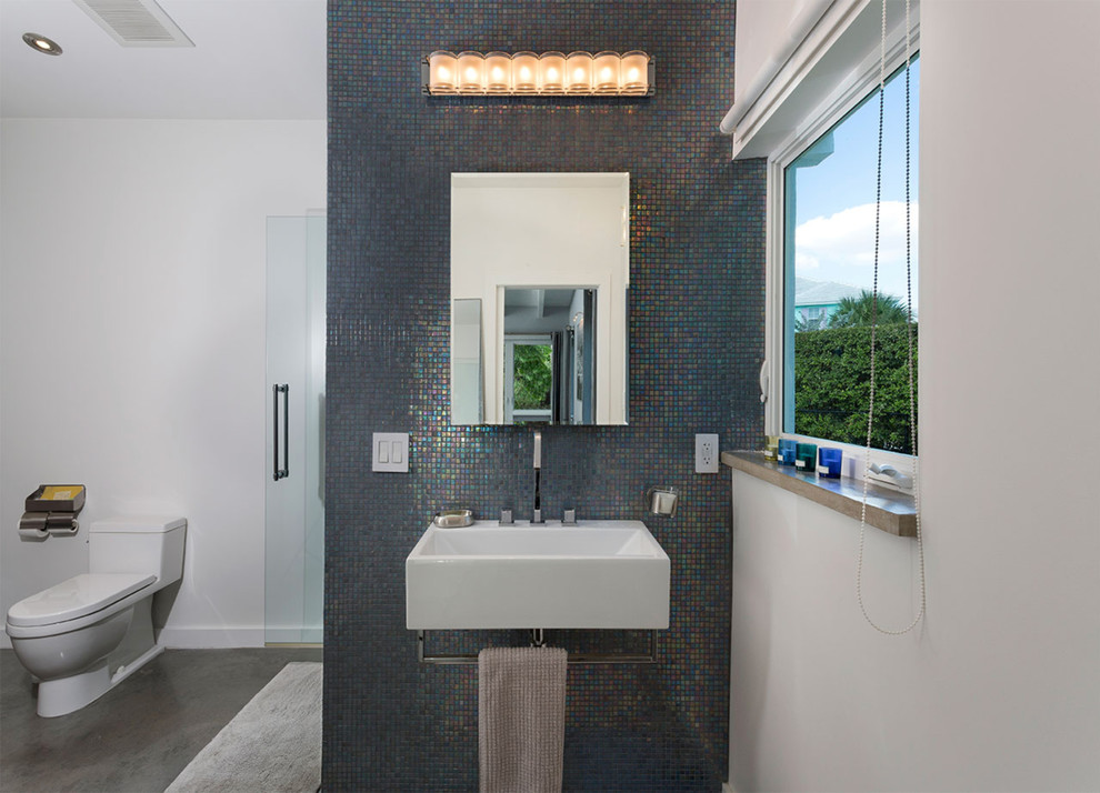 Medium sized midcentury ensuite bathroom in Miami with an alcove shower, a one-piece toilet, multi-coloured tiles, glass tiles, white walls, concrete flooring, a wall-mounted sink, solid surface worktops, grey floors, a hinged door and white worktops.