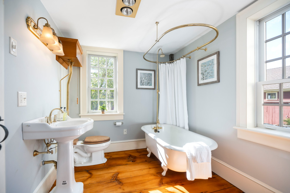 Rural bathroom in Boston with a claw-foot bath, a shower/bath combination, grey walls, medium hardwood flooring, a pedestal sink, brown floors, a shower curtain and a single sink.