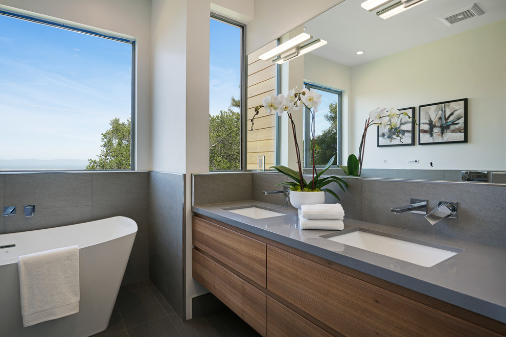Contemporary bathroom in San Francisco with flat-panel cabinets, medium wood cabinets, a freestanding bath, grey tiles, white walls, a submerged sink, grey floors and grey worktops.