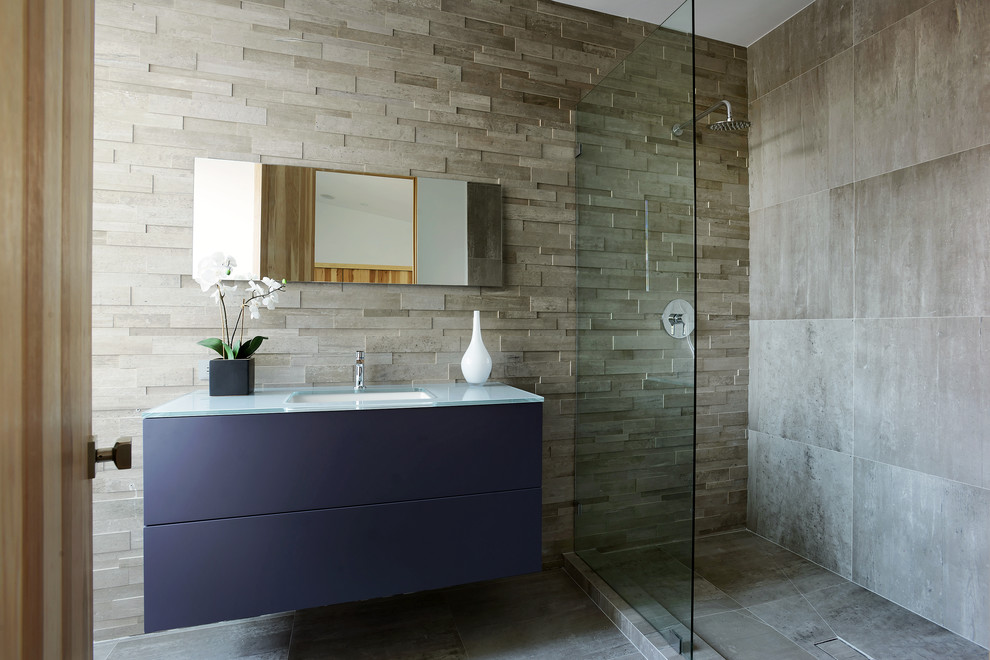 Modelo de cuarto de baño rectangular actual con lavabo bajoencimera, armarios con paneles lisos, puertas de armario azules, baldosas y/o azulejos grises, aseo y ducha y encimera de vidrio