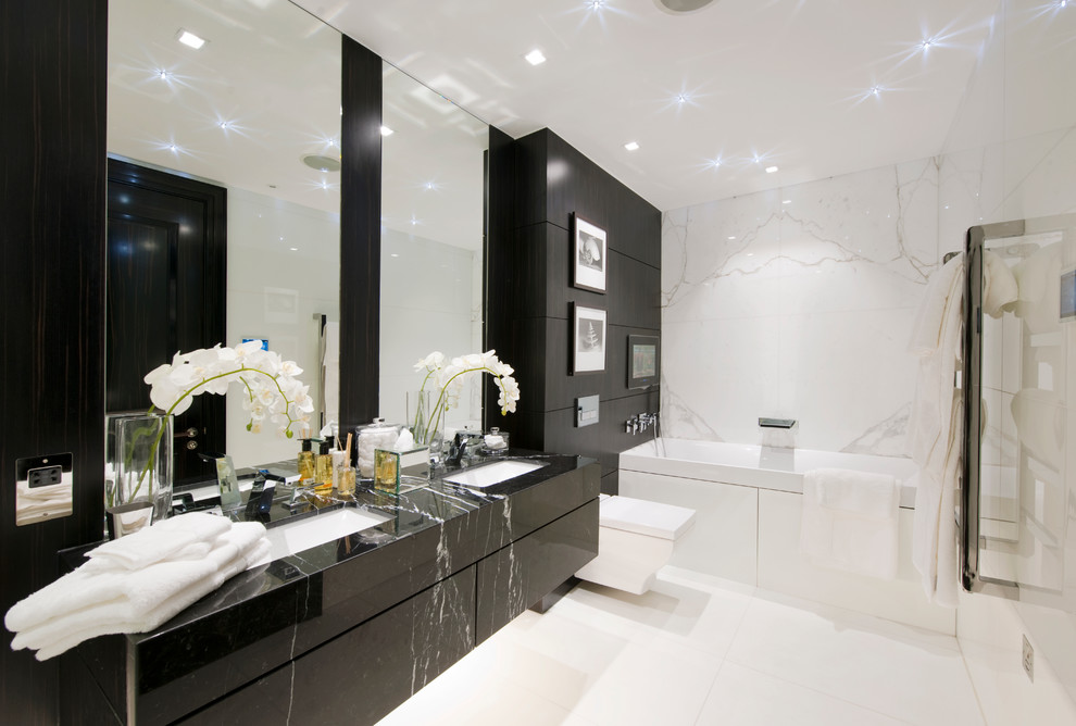 Diseño de cuarto de baño actual con lavabo bajoencimera, armarios con paneles lisos, puertas de armario negras, bañera empotrada, sanitario de pared, baldosas y/o azulejos blancos y encimeras negras