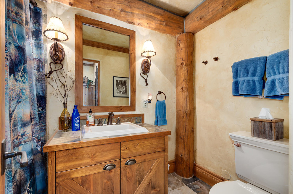 Modelo de cuarto de baño rural con puertas de armario de madera oscura, ducha empotrada, paredes beige, lavabo encastrado, encimera de azulejos, suelo gris, ducha con cortina y encimeras marrones
