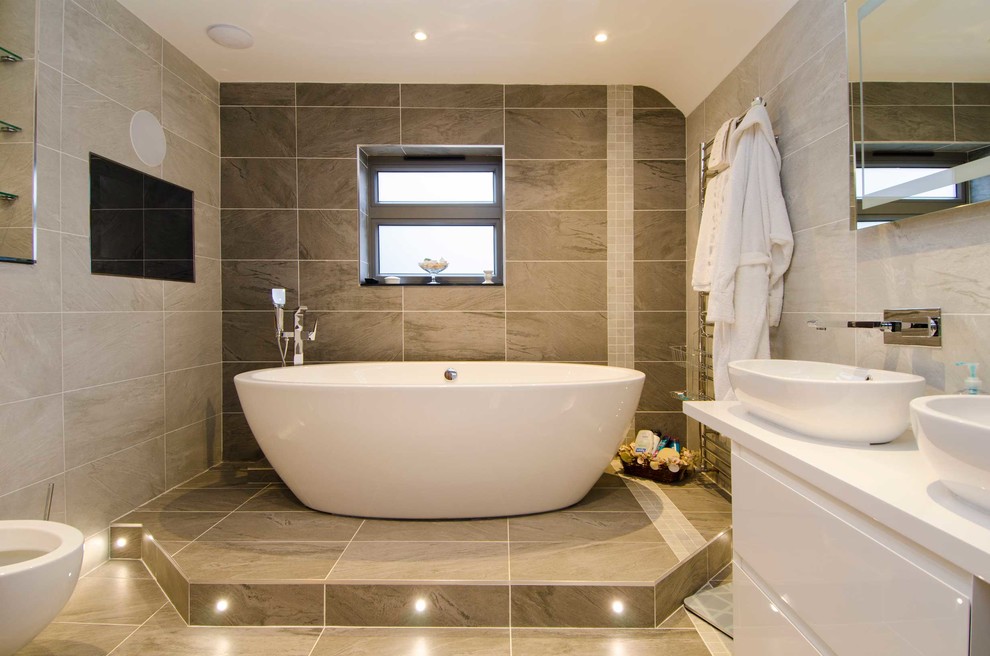 Cette image montre une salle de bain design avec une vasque, un placard à porte plane, des portes de placard blanches, une baignoire indépendante, un carrelage beige et un mur beige.