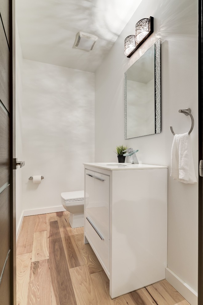 Cette image montre une salle d'eau design de taille moyenne avec un placard à porte plane, des portes de placard blanches, WC séparés, un mur blanc, parquet clair, un lavabo encastré et un plan de toilette en surface solide.
