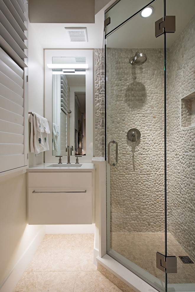 Cette image montre une salle de bain marine avec un placard à porte plane, des portes de placard blanches, une douche d'angle, un carrelage gris, une plaque de galets, un mur blanc, un sol beige, une cabine de douche à porte battante et un plan de toilette blanc.