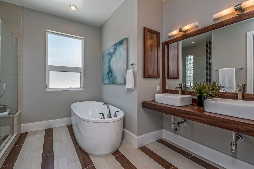 Foto de cuarto de baño principal actual grande con bañera exenta, paredes grises, suelo de baldosas de porcelana, lavabo sobreencimera y encimera de madera