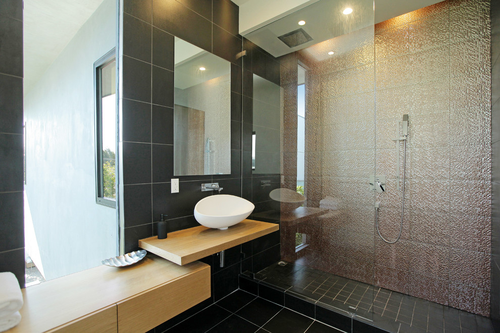 Modern inredning av ett badrum, med ett fristående handfat, släta luckor, skåp i mellenmörkt trä, träbänkskiva, en dusch i en alkov och svart kakel