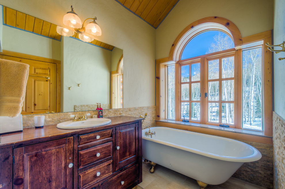 This is an example of a medium sized rustic ensuite bathroom in Denver with a built-in sink, raised-panel cabinets, dark wood cabinets, wooden worktops, a claw-foot bath, beige tiles, glass tiles, yellow walls and ceramic flooring.