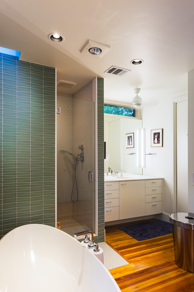 Réalisation d'une douche en alcôve design avec un placard à porte plane, des portes de placard blanches, un carrelage vert, une baignoire indépendante, un carrelage en pâte de verre, un sol en bois brun, un lavabo encastré et une fenêtre.