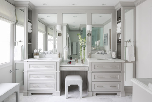 Elegant light gray bathroom cabinets with double sinks, marble countertop, built-in vanity desk, and large mirrors in a bright luxury bathroom
