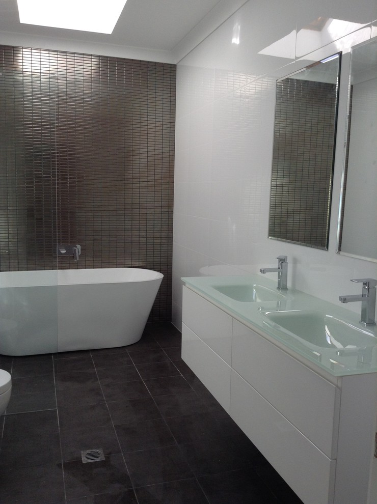 На фото: главная ванная комната среднего размера в стиле модернизм с монолитной раковиной, плоскими фасадами, белыми фасадами, стеклянной столешницей, отдельно стоящей ванной, открытым душем, коричневой плиткой, керамической плиткой и полом из керамической плитки с