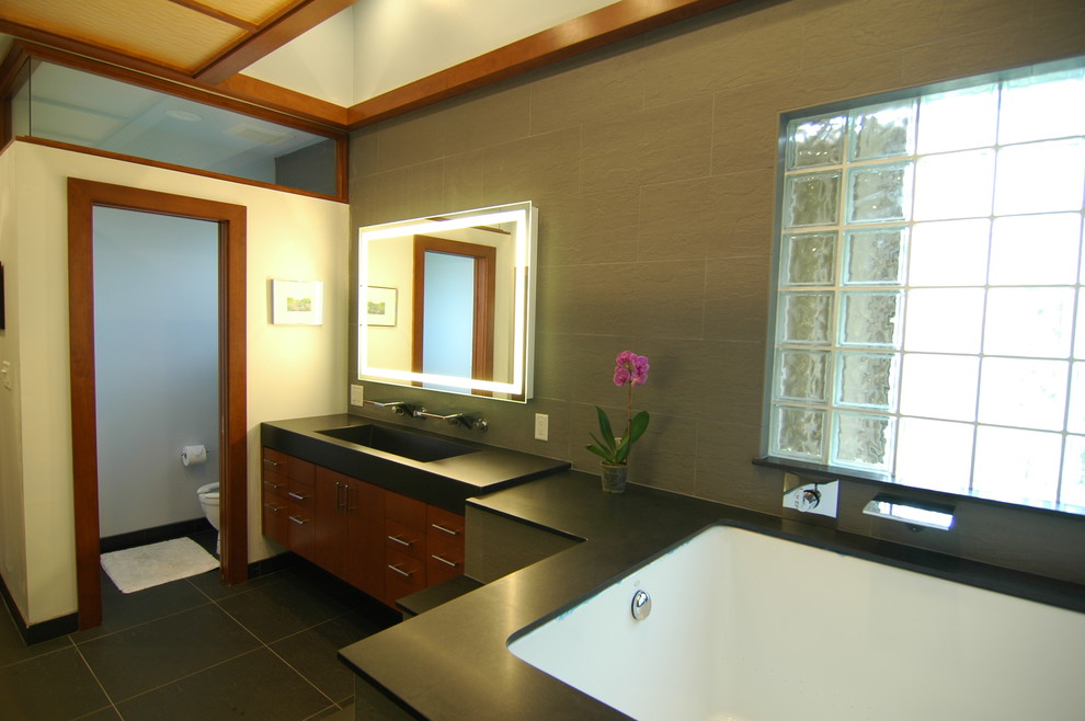 Immagine di una stanza da bagno etnica con lavabo rettangolare, ante lisce, ante in legno scuro, vasca sottopiano, piastrelle nere e piastrelle in pietra