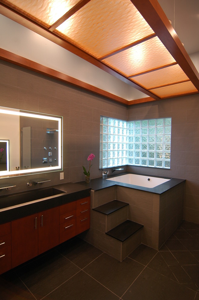 Пример оригинального дизайна: ванная комната в восточном стиле с раковиной с несколькими смесителями, плоскими фасадами, фасадами цвета дерева среднего тона, полновстраиваемой ванной, открытым душем, черной плиткой и каменной плиткой