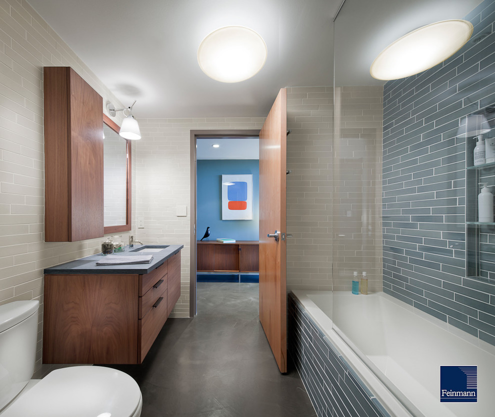 Modelo de cuarto de baño actual con lavabo bajoencimera, armarios con paneles lisos, puertas de armario de madera en tonos medios, bañera encastrada, combinación de ducha y bañera, sanitario de dos piezas y baldosas y/o azulejos grises
