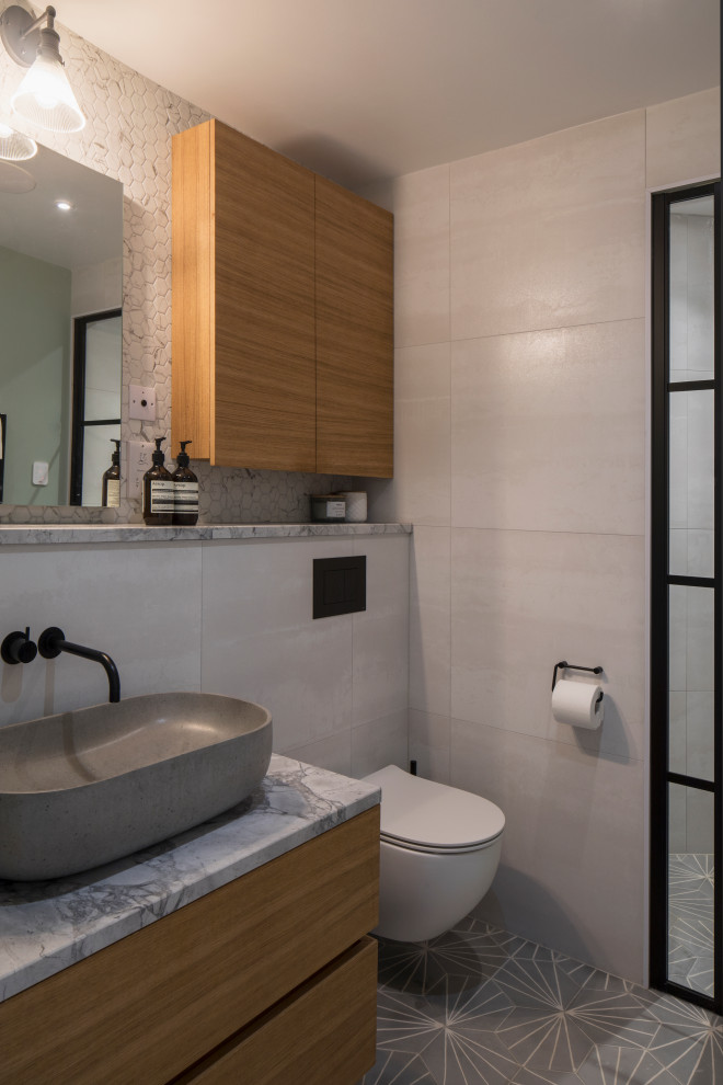 Bild på ett mellanstort funkis grå grått badrum, med släta luckor, skåp i mellenmörkt trä, en dusch i en alkov, en vägghängd toalettstol, grå kakel, ett fristående handfat och grått golv