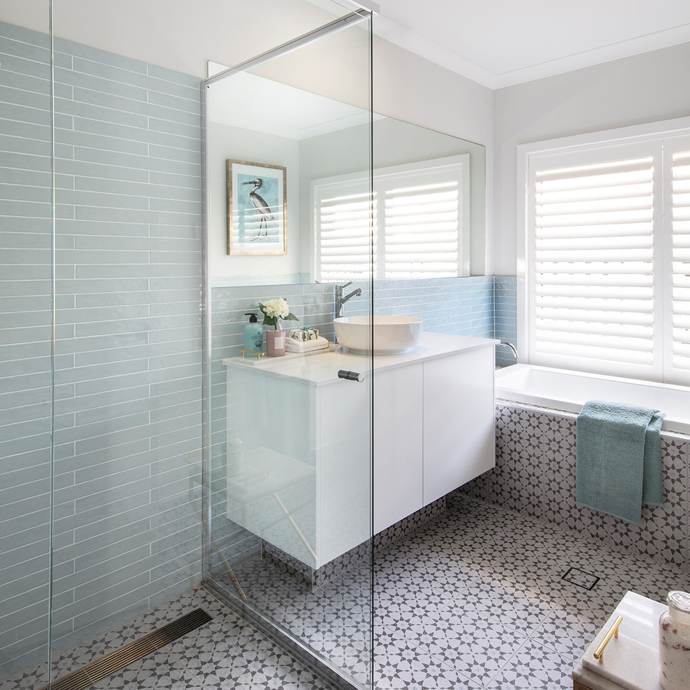 Modern inredning av ett vit vitt badrum, med släta luckor, vita skåp, ett platsbyggt badkar, en kantlös dusch, blå kakel, glaskakel, grå väggar, ett fristående handfat, flerfärgat golv och med dusch som är öppen