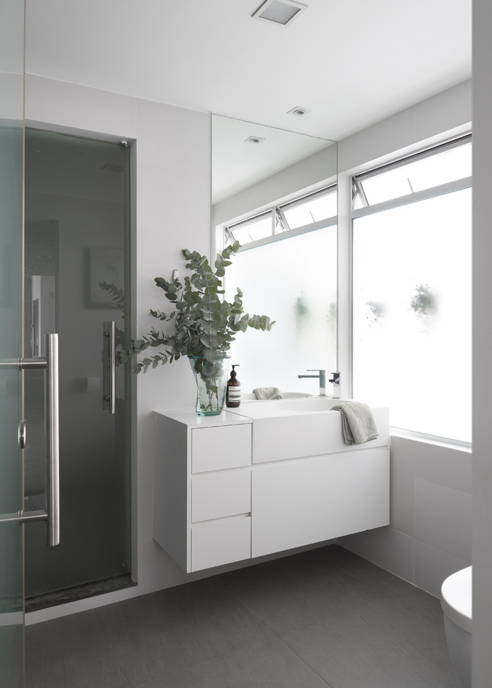Cette photo montre une salle de bain industrielle avec un placard à porte plane, des portes de placard blanches, WC suspendus, un mur gris, un plan vasque, un sol gris, une cabine de douche à porte battante et un plan de toilette blanc.