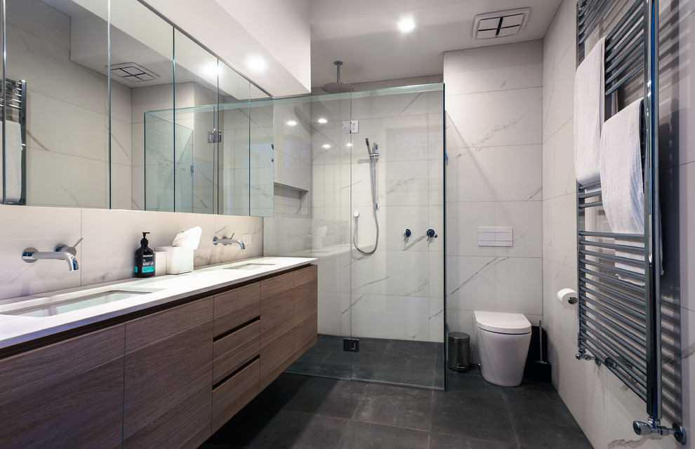 Medium sized modern ensuite bathroom in Melbourne with flat-panel cabinets, medium wood cabinets, a wall mounted toilet, grey tiles, porcelain tiles, white walls, porcelain flooring, a submerged sink, engineered stone worktops and a corner shower.