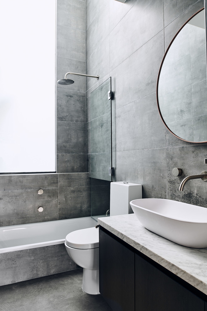 Idée de décoration pour une salle de bain grise et noire design avec un placard à porte plane, des portes de placard noires, une baignoire posée, un combiné douche/baignoire, un carrelage gris, un mur gris, sol en béton ciré, une vasque, un sol gris et un plan de toilette blanc.