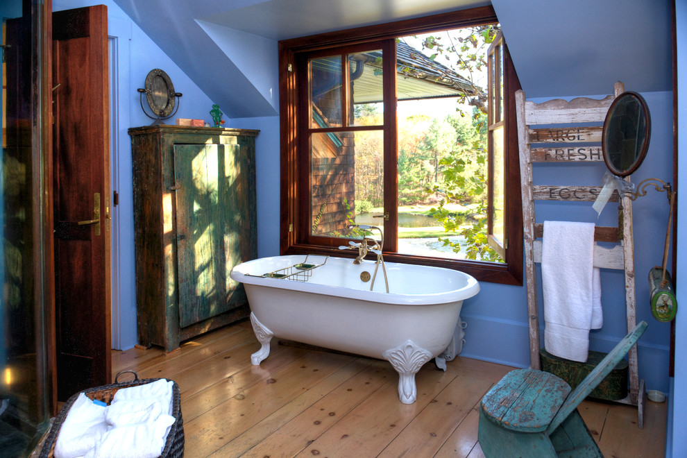 Bild på ett rustikt badrum, med skåp i slitet trä, ett badkar med tassar, ljust trägolv och blå väggar