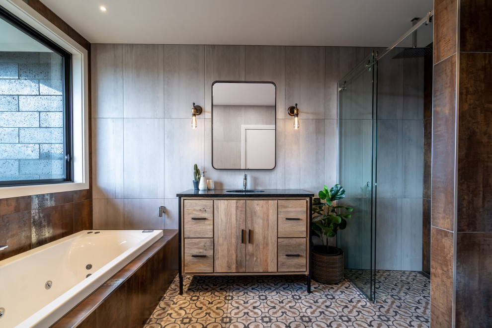 Cette image montre une salle de bain principale urbaine en bois brun de taille moyenne avec un plan de toilette en granite, un plan de toilette noir, une baignoire posée, un carrelage gris, un lavabo encastré, un sol multicolore et un placard à porte plane.