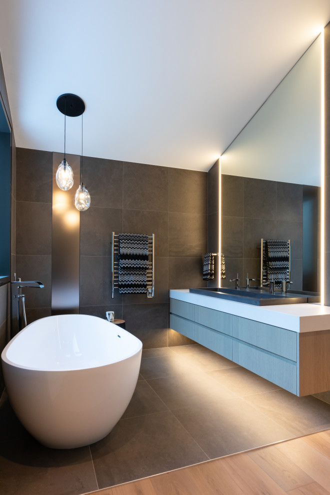 Design ideas for a large contemporary ensuite bathroom in Other with flat-panel cabinets, light wood cabinets, a freestanding bath, a double shower, a wall mounted toilet, grey tiles, porcelain tiles, white walls, porcelain flooring, a trough sink, engineered stone worktops, grey floors, an open shower, white worktops, an enclosed toilet, double sinks and a floating vanity unit.