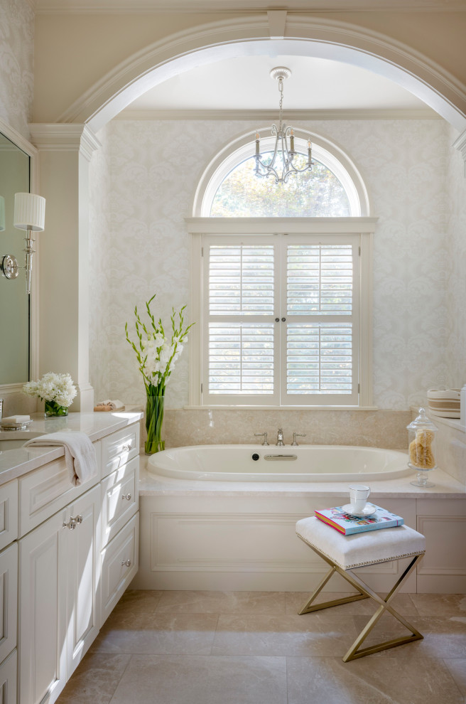 Klassisk inredning av ett vit vitt badrum, med luckor med upphöjd panel, vita skåp, ett platsbyggt badkar, beige kakel, stenhäll, beige väggar, ett undermonterad handfat och beiget golv