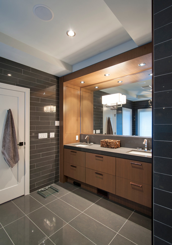 Modelo de cuarto de baño actual con lavabo bajoencimera, armarios con paneles lisos, puertas de armario de madera oscura, baldosas y/o azulejos grises, baldosas y/o azulejos de vidrio y encimeras grises