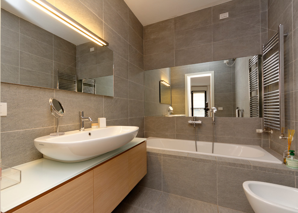Immagine di una stanza da bagno padronale contemporanea di medie dimensioni con lavabo a consolle, top in vetro, vasca da incasso, WC sospeso, piastrelle grigie, piastrelle in pietra e pareti grigie