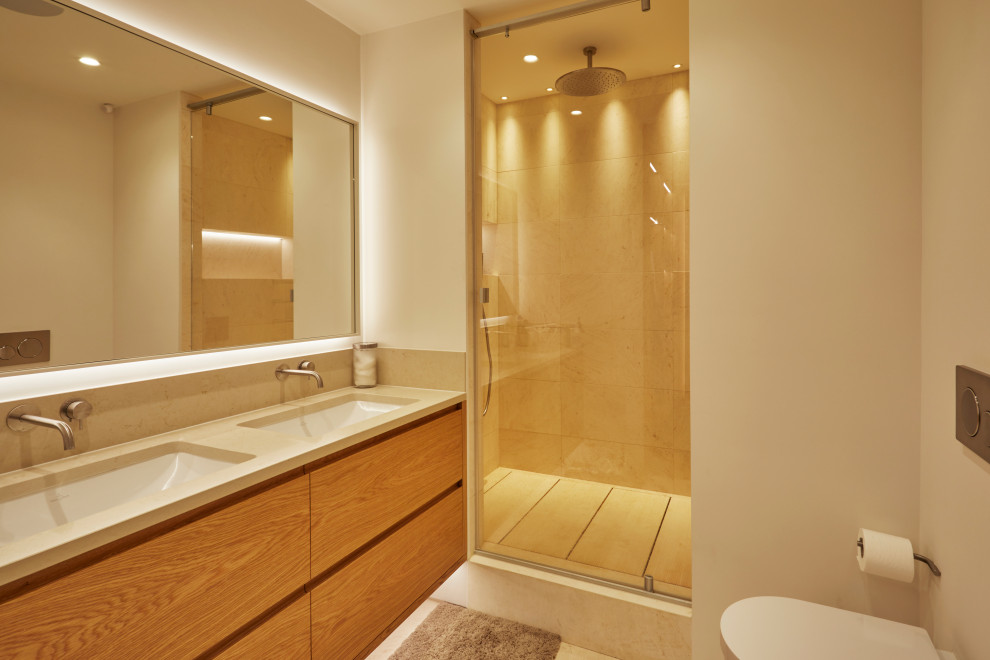 Kids' beige tile and porcelain tile ceramic tile and beige floor doorless shower photo in London with flat-panel cabinets, medium tone wood cabinets, a wall-mount toilet, beige walls, an undermount sink, limestone countertops, a hinged shower door and beige countertops