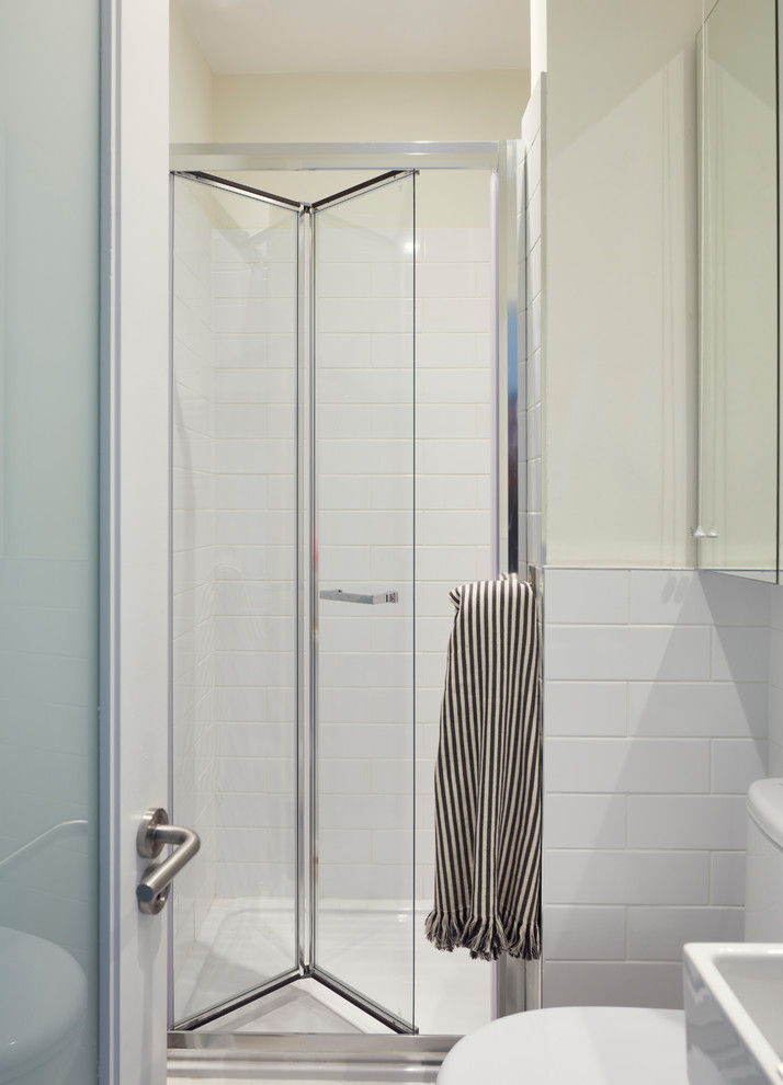 Bild på ett litet nordiskt badrum med dusch, med luckor med glaspanel, vita skåp, en dubbeldusch, en toalettstol med hel cisternkåpa, vit kakel, keramikplattor, vita väggar, vinylgolv, ett piedestal handfat, vitt golv och dusch med gångjärnsdörr