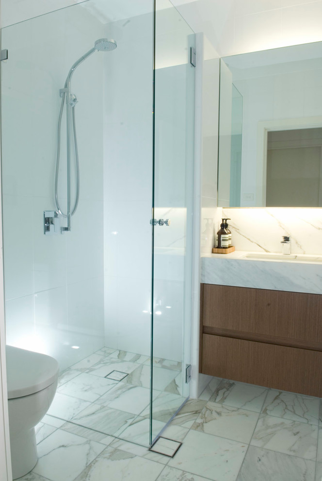 Modern inredning av ett litet badrum med dusch, med ett undermonterad handfat, skåp i mellenmörkt trä, marmorbänkskiva, en hörndusch, en toalettstol med hel cisternkåpa, vit kakel, porslinskakel, vita väggar och marmorgolv