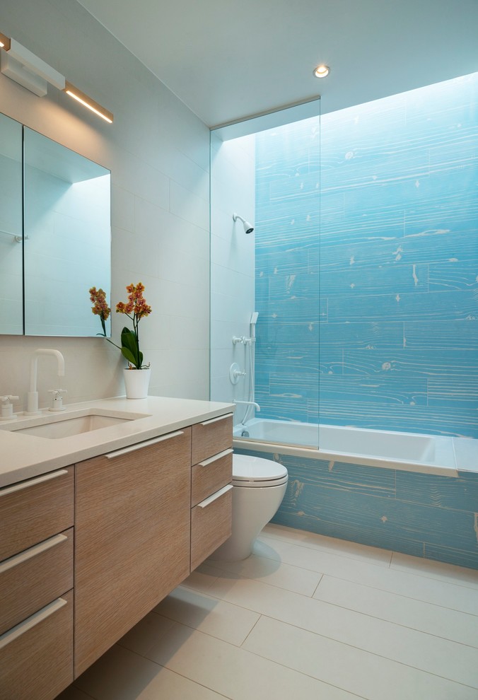 Imagen de cuarto de baño contemporáneo con armarios con paneles lisos, puertas de armario de madera clara, bañera encastrada, combinación de ducha y bañera, baldosas y/o azulejos azules, paredes grises, lavabo bajoencimera, suelo gris y encimeras blancas