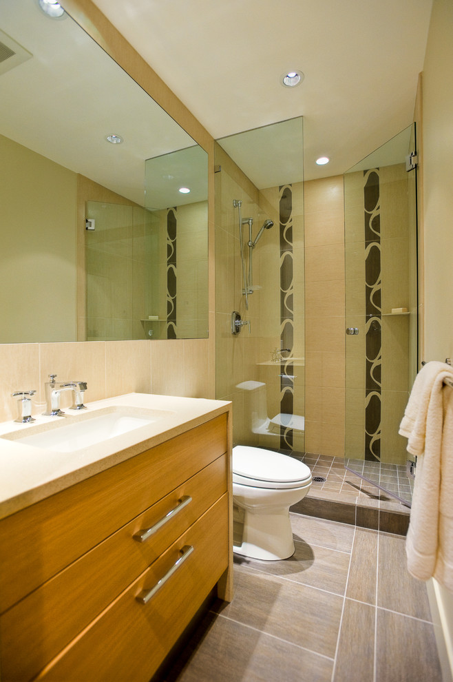 Inredning av ett modernt badrum, med ett undermonterad handfat, släta luckor, skåp i mellenmörkt trä, en dusch i en alkov, en toalettstol med hel cisternkåpa och beige kakel