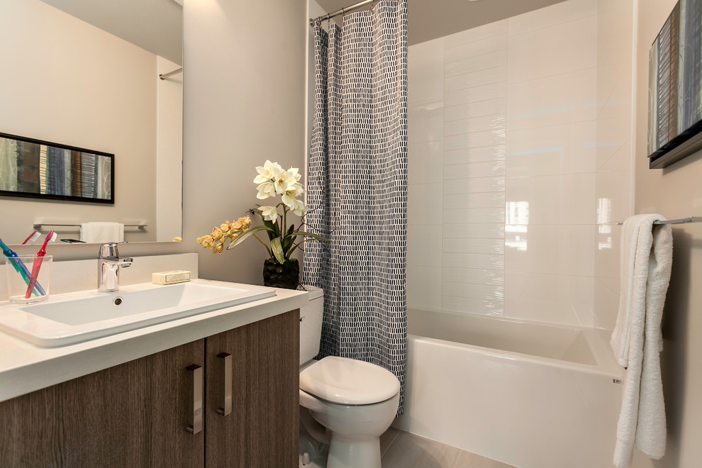 This is an example of a small contemporary bathroom in Vancouver with a built-in sink, flat-panel cabinets, medium wood cabinets, engineered stone worktops, an alcove bath, a shower/bath combination, a two-piece toilet, white tiles, porcelain tiles, grey walls, porcelain flooring and a shower curtain.
