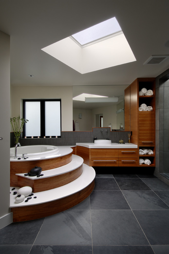 Modern inredning av ett badrum, med ett fristående handfat, släta luckor, skåp i mellenmörkt trä, ett platsbyggt badkar och svart kakel