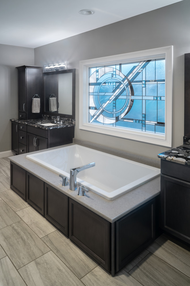 Example of a mid-sized trendy master porcelain tile and beige floor drop-in bathtub design in St Louis with flat-panel cabinets, dark wood cabinets, gray walls, an undermount sink, granite countertops and black countertops
