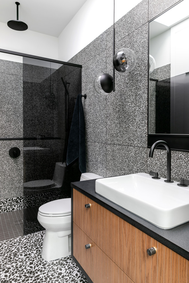 Пример оригинального дизайна: маленькая главная ванная комната в современном стиле с плоскими фасадами, душем в нише, унитазом-моноблоком, черно-белой плиткой, керамогранитной плиткой, белыми стенами, настольной раковиной, столешницей из гранита, черным полом, душем с распашными дверями, черной столешницей, тумбой под одну раковину, подвесной тумбой, фасадами цвета дерева среднего тона и полом из терраццо для на участке и в саду